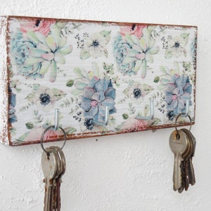 May include: A wooden key holder with a floral design featuring succulents and flowers in shades of pink, blue, and green. The holder has five hooks for hanging keys.