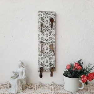 Könnte beinhalten: Ein weißer Schlüsselhalter mit floralem Muster und fünf Haken. Der Schlüsselhalter ist an einer weißen Wand montiert. An den Haken hängen fünf Schlüssel.
