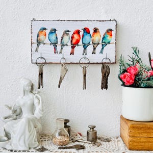 May include: A white wooden key holder with a painted design of colorful birds. The holder has five hooks for hanging keys.