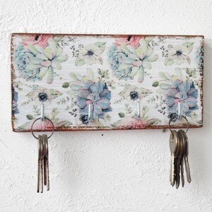 May include: A wooden key holder with a floral design featuring succulents and flowers in shades of pink, green, and blue. The holder has five hooks for hanging keys.