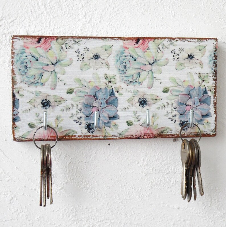 May include: A wooden key holder with a floral design featuring succulents and flowers in shades of pink, green, and blue. The holder has five hooks for hanging keys.