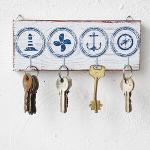 K&ouml;nnte beinhalten: Ein wei&szlig;er Holz-Schl&uuml;sselhalter mit blauen nautischen Designs. Der Halter hat vier Haken, an denen Schl&uuml;ssel h&auml;ngen. Die Designs beinhalten einen Leuchtturm, eine Schiffsschraube, einen Anker und einen Kompass.