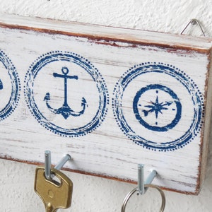 K&ouml;nnte beinhalten: Wei&szlig;er Holzschl&uuml;sselhalter mit drei Haken. Der Halter hat ein nautisches Thema mit blauen und wei&szlig;en Designs eines Propellers, eines Ankers und eines Kompasses.