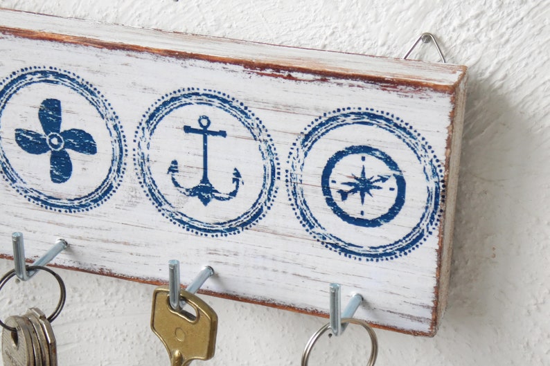 K&ouml;nnte beinhalten: Wei&szlig;er Holzschl&uuml;sselhalter mit drei Haken. Der Halter hat ein nautisches Thema mit blauen und wei&szlig;en Designs eines Propellers, eines Ankers und eines Kompasses.