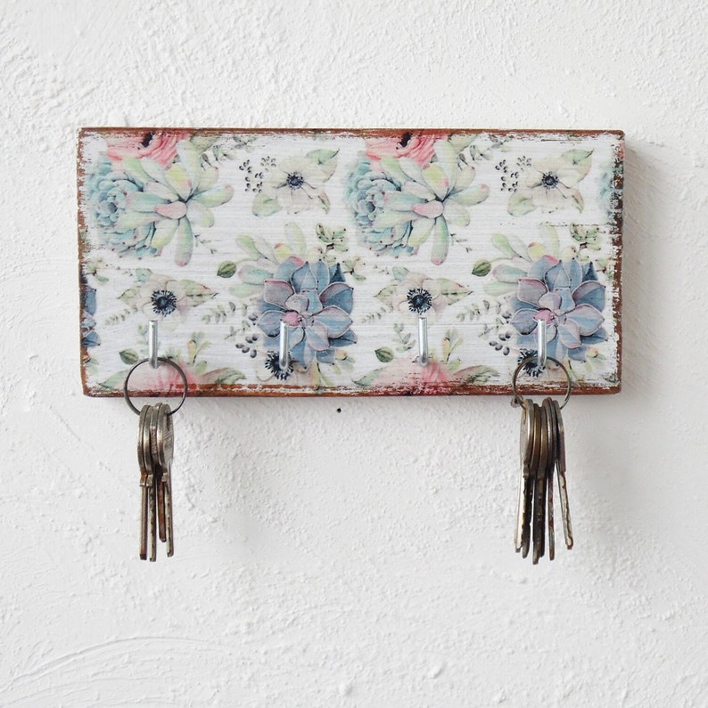 A rectangular wooden key holder featuring a pattern of pastel succulents and flowers on a white background. The surface of the key holder shows light distressing, and the brown wood edges are visible. Four metal hooks are present on the key holder, two of which are holding bunches of keys.