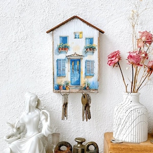 Könnte beinhalten: Ein hölzerner Schlüsselhalter in Form eines Hauses mit einer blauen Tür und weißen Fensterläden. Das Haus ist im Aquarellstil bemalt und hat Blumenkästen an den Fenstern. An Haken an der Unterseite des Hauses hängen Schlüssel.