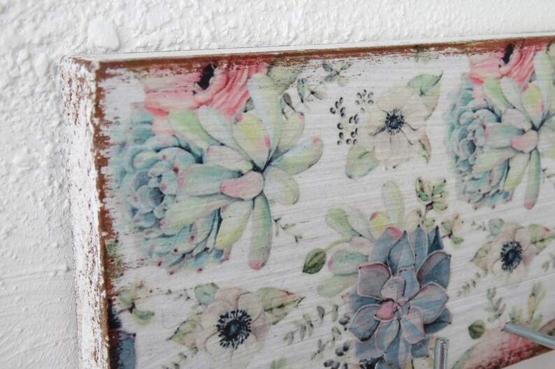 May include: A wooden wall-mounted key holder with a floral design featuring succulents and anemones in shades of pink, blue, and green. The wood has a distressed white finish.