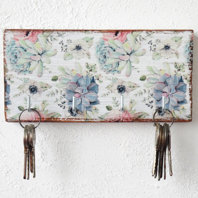 May include: A white wooden key holder with a floral design featuring succulents and flowers in shades of pink, green, and blue. The key holder has three hooks and is distressed for a vintage look.