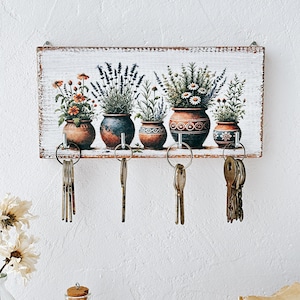 May include: White wooden key holder with a painted design of five flower pots with different flowers. The key holder has five hooks for hanging keys.