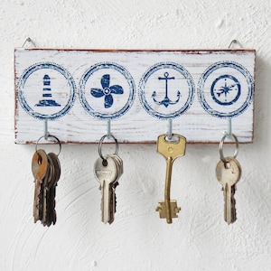 May include: White wooden key holder with five hooks. The key holder has a nautical theme with blue and white circles featuring a lighthouse, compass, anchor, and propeller. Five keys are hanging on the hooks.