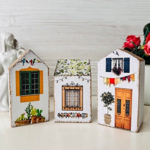 Pode incluir: Três casas de madeira em miniatura com detalhes pintados. Cada casa tem um design diferente, incluindo uma janela com persianas, uma porta com uma coroa e um telhado frondoso. As casas são pintadas de branco com detalhes coloridos.