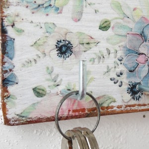 May include: A white wooden key holder with a floral design and two metal hooks. The hooks are silver and have a ring attached to one of them.