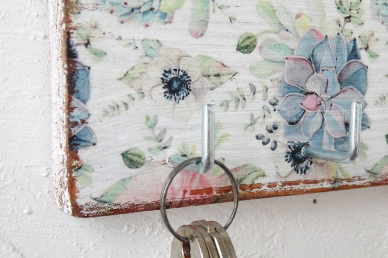 May include: A white wooden key holder with a floral design and two metal hooks. The hooks are silver and have a ring attached to one of them.
