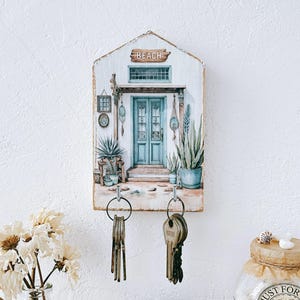 May include: A wooden key holder with a painted beach scene featuring a blue door, plants, and the word "BEACH" on a sign. Two sets of keys hang from metal hooks below the artwork. The key holder is mounted on a white wall.