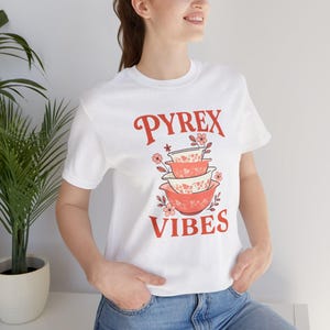 May include: White t-shirt with a retro design featuring the text "PYREX VIBES" in red script above a stack of vintage Pyrex bowls in shades of pink and white. The bowls are decorated with floral patterns.