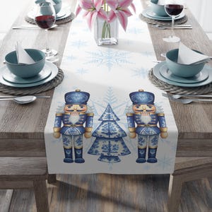 May include: A white table runner featuring a repeating pattern of two blue nutcrackers flanking a blue and white patterned Christmas tree, accented with light blue snowflakes. The design is printed on a long fabric strip for table decor.
