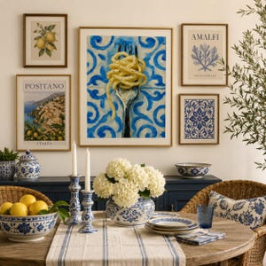 May include: A dining room scene featuring a gallery wall with five framed prints, including a central illustration of spaghetti on a fork against blue tiles, flanked by lemon, coastal, and floral art, with a table set with white flowers.