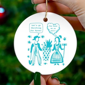 May include: A white ceramic ornament with a teal illustration and gold string. The illustration depicts two figures with text bubbles that read "Let's go Thrifting for Pyrex!" and "You know me so well!"