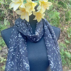May include: A black chiffon scarf with a white musical note print. The scarf is draped around a mannequin's neck and has a loose, flowing style.