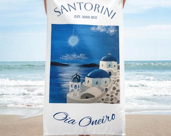 May include: White beach towel with a blue and white illustration of buildings in Santorini, Greece. The text "Santorini Est. 3000 BCE" and "Oia Oneiro" are printed on the towel.