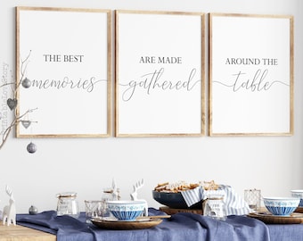May include: Three framed prints with the text "The best memories are made gathered around the table" in a modern calligraphy font. The prints are hanging on a white wall above a wooden table set with blue and white dishes and a blue tablecloth.