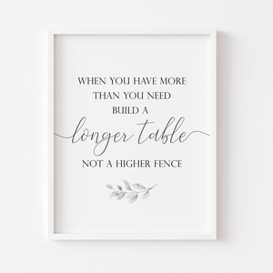 May include: A white framed print with a quote that reads "When you have more than you need build a longer table not a higher fence". The quote is in black text with a decorative script font. A small green watercolour leaf design is at the bottom of the print.
