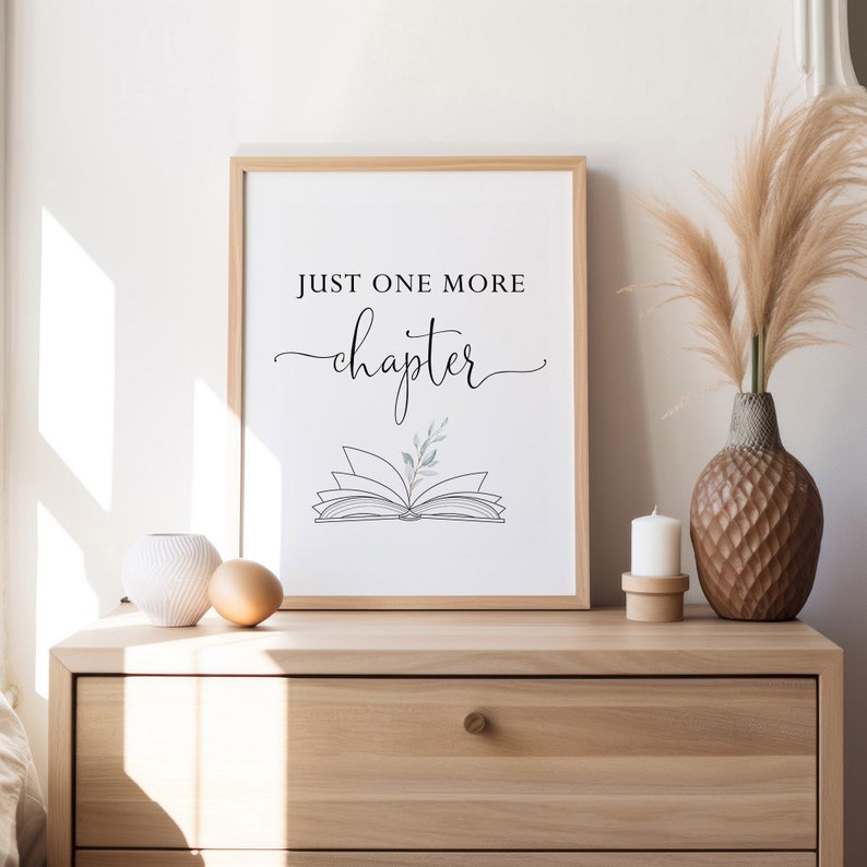 May include: A framed print with the text "Just one more chapter" in black script font. A stylized drawing of an open book with a small sprig of leaves on top is below the text. The print is on a white background and is framed in a light wood frame.