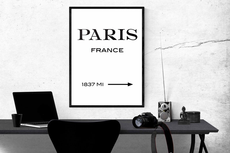 Op de afbeelding: Zwart-witte print met de tekst "PARIS FRANCE 1837 MI" en een pijl die naar rechts wijst. De print is ingelijst en hangt aan een witte muur.