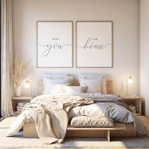 Puede incluir: Dos impresiones enmarcadas con el texto "With you" y "I am home" en una fuente cursiva. Las impresiones están colgadas sobre una cama con ropa de cama blanca y una cabecera de madera.