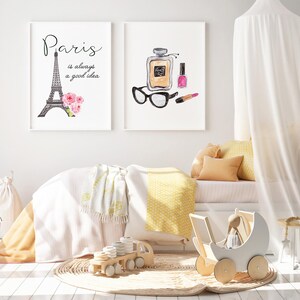 May include: A white canopy bed with yellow and white bedding, a white blanket, and a white doll stroller. Two framed prints hang on the wall above the bed. One print features a black and white illustration of the Eiffel Tower with the text "Paris is always a good idea". The other print features a watercolor illustration of a perfume bottle, lipstick, and sunglasses.