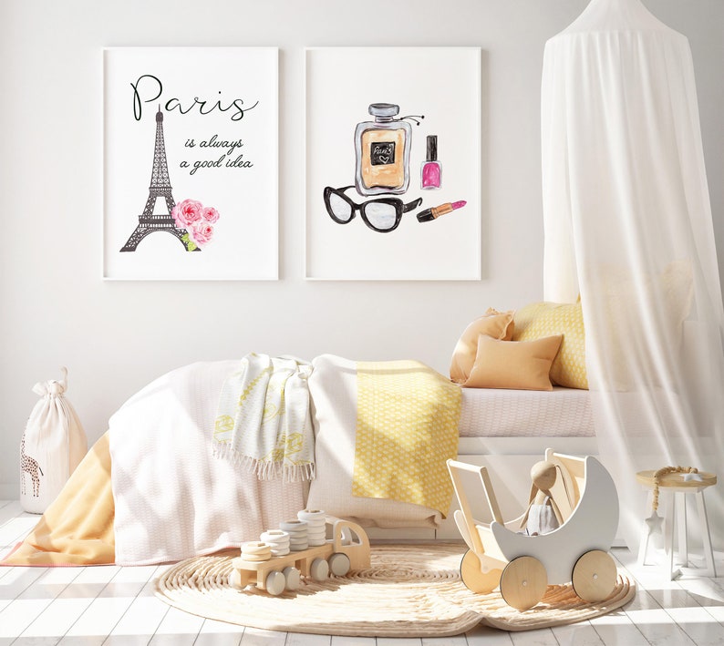 May include: A white canopy bed with yellow and white bedding, a white blanket, and a white doll stroller. Two framed prints hang on the wall above the bed. One print features a black and white illustration of the Eiffel Tower with the text "Paris is always a good idea". The other print features a watercolor illustration of a perfume bottle, lipstick, and sunglasses.