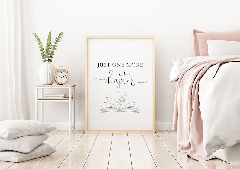 May include: A black and white printable wall art print with the text "Just one more chapter" and a line drawing of an open book with a small sprig of leaves on top. The print is framed in a gold frame and is hanging on a white wall.