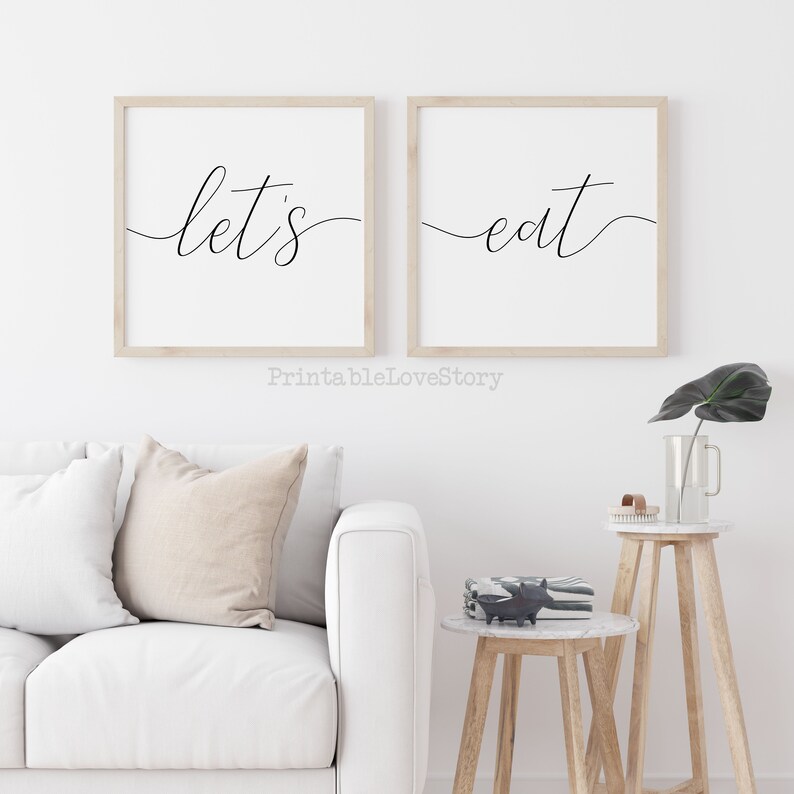 May include: Two framed prints with the text "let's eat" in a modern calligraphy style. The prints are hung on a white wall above a white couch and two wooden side tables.