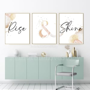 Può includere: Tre stampe d'arte da parete stampabili con disegni ad acquerello dorati e rosa. Le stampe presentano le parole "Rise", "&" e "Shine" in calligrafia nera.