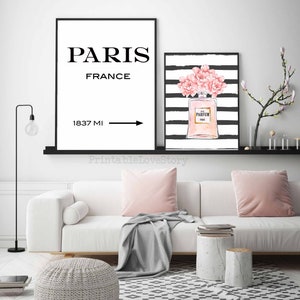 Op de afbeelding: Twee zwart ingelijste prints op een witte muur boven een witte bank. De linker print zegt "Parijs, Frankrijk" met een pijl naar rechts en "1837 MI". De rechter print heeft zwart-witte strepen met een roze parfumfles en roze bloemen.