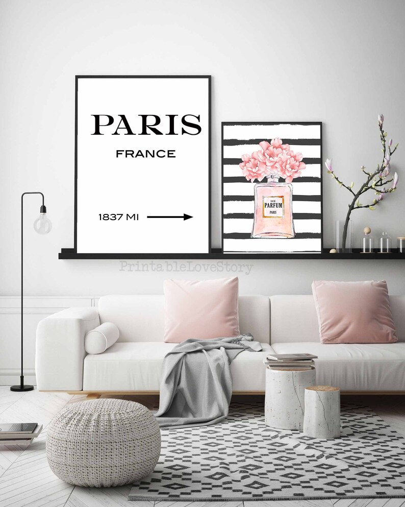 Op de afbeelding: Twee zwart ingelijste prints op een witte muur boven een witte bank. De linker print zegt "Parijs, Frankrijk" met een pijl naar rechts en "1837 MI". De rechter print heeft zwart-witte strepen met een roze parfumfles en roze bloemen.
