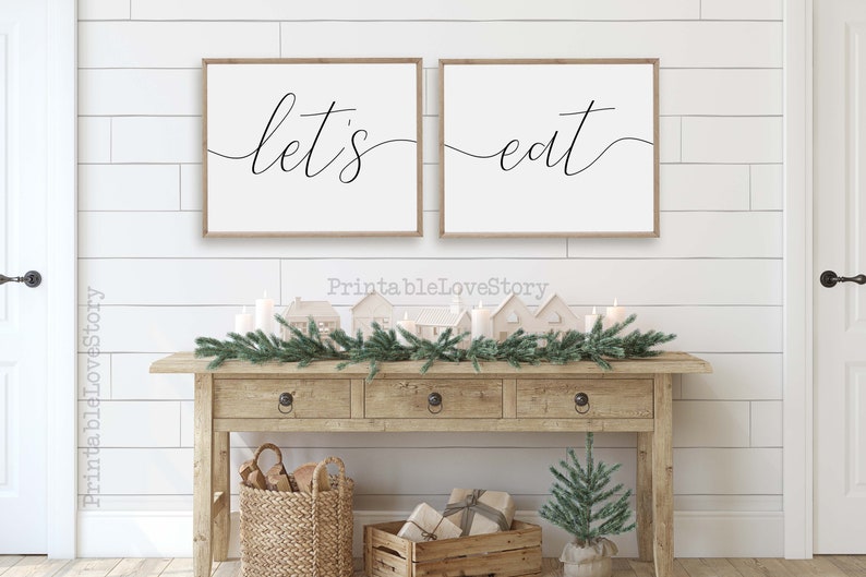 May include: Two white framed signs with black calligraphy on a white wood background. The signs read "let's" and "eat". The signs are hanging above a wooden console table with a green garland, candles, and small white houses.
