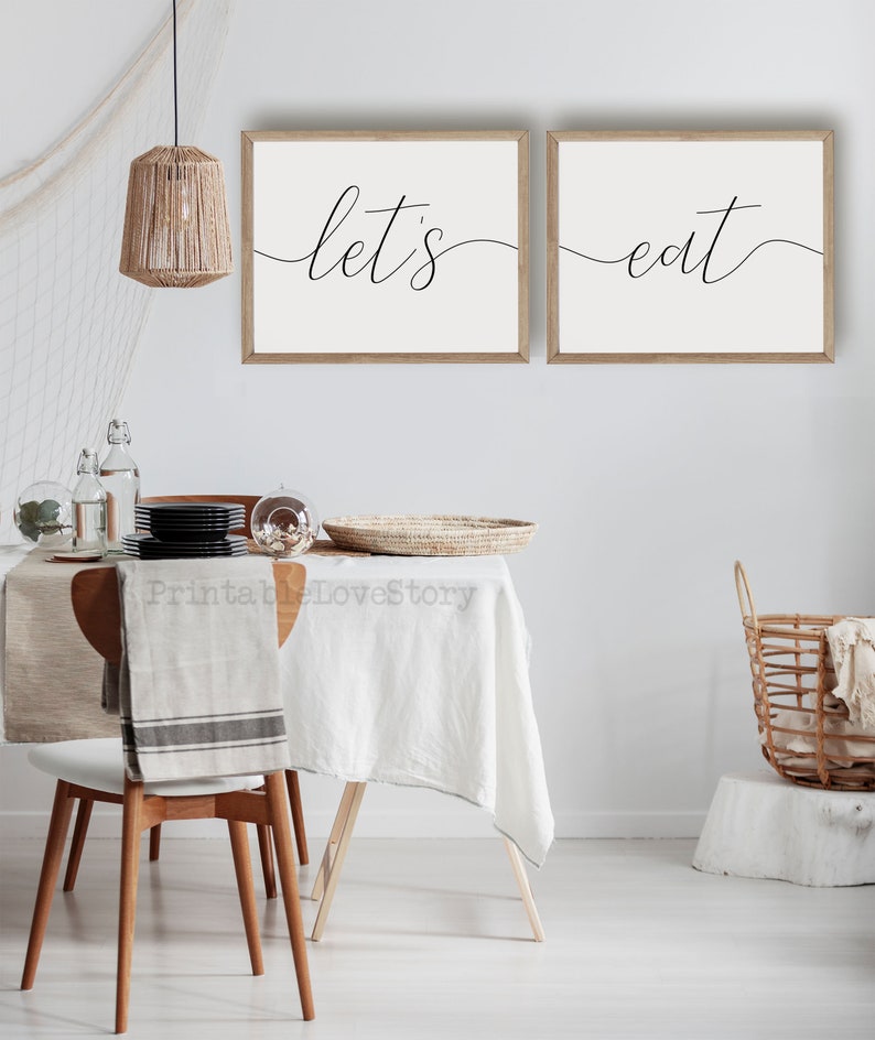 May include: Two framed prints with the text "let's eat" in a modern calligraphy style. The prints are hanging on a white wall above a dining table with a white tablecloth and a wicker basket.