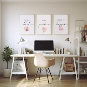 May include: Three framed motivational posters with pink watercolor flowers and black calligraphy text. The posters read "Work hard", "Stay positive", and "Make it happen". The posters are hanging on a white wall above a white desk with a computer, a chair, and a lamp.