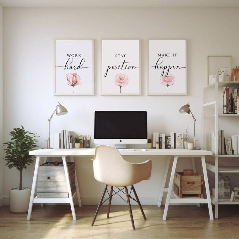 May include: Three framed motivational posters with pink watercolor flowers and black calligraphy text. The posters read "Work hard", "Stay positive", and "Make it happen". The posters are hanging on a white wall above a white desk with a computer, a chair, and a lamp.