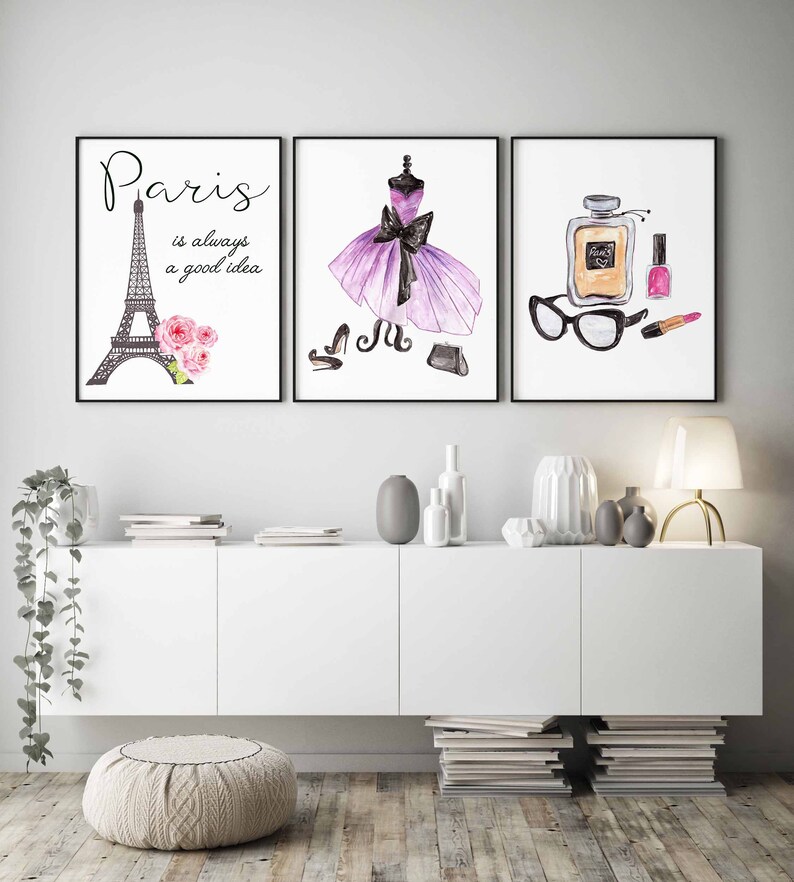 May include: Three framed watercolor prints with a Parisian theme. The first print features a black and white illustration of the Eiffel Tower with pink flowers. The second print features a purple dress with a black bow and black shoes. The third print features a bottle of perfume, a pair of glasses, a lipstick, and a nail polish bottle.