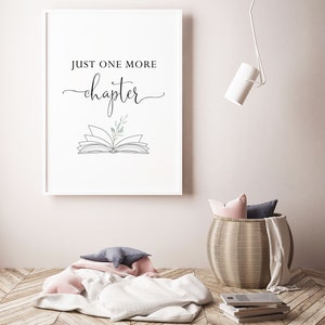 May include: A white framed print with the text "Just one more chapter" and a line drawing of an open book with a small sprig of greenery on top. The print is hanging on a white wall above a wicker basket with a pink and gray throw blanket.