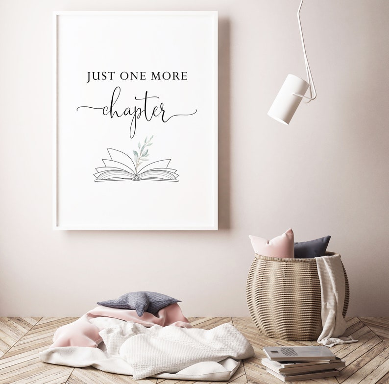 May include: A white framed print with the text "Just one more chapter" and a line drawing of an open book with a small sprig of greenery on top. The print is hanging on a white wall above a wicker basket with a pink and gray throw blanket.