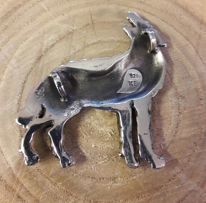 May include: Silver pendant in the shape of a wolf howling at the moon. The pendant has a detailed design and is marked "925 KF" on the back.