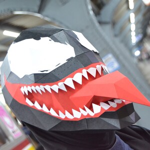 以下が含まれることがあります： 黒い頭部、大きな白い目、広い赤い口、鋭い白い歯、そして突き出た長い赤い舌が特徴のローポリゴンペーパークラフトマスク。このマスクは幾何学的なファセットデザインで、角にはBAMBOOGILAというロゴが記載されています。