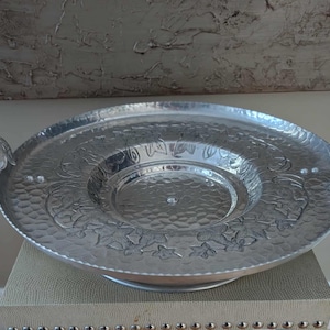 May include: A silver metal serving tray with two handles. The tray has a hammered texture and embossed floral designs around the rim. The handles are shaped like leaves.