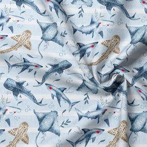 May include: Fabric featuring a repeating pattern of blue sharks, stingrays, and spotted tan fish on a light blue and white striped background. The marine animals are illustrated with small red dots for eyes and interspersed with gray leafy sprigs.