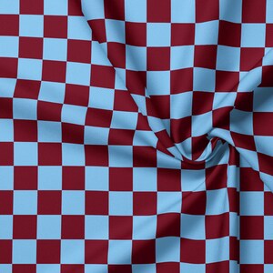 Op de afbeelding: Een stof met een herhalend ruitpatroon van lichtblauwe en donkerbordeauxrode vierkanten. Het materiaal is gedrapeerd in zachte plooien, wat het consistente geometrische rasterontwerp over het gehele oppervlak van het textiel benadrukt.