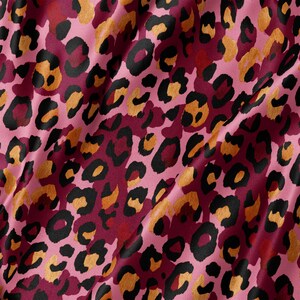 May include: A fabric with a repeating leopard print pattern featuring black outlines, golden-brown centres, and a deep magenta background. The material has a smooth, slightly reflective surface with soft folds creating subtle shadows across the textile.