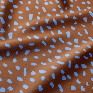 May include: A brown fabric featuring a repeating pattern of light blue, irregular, rounded spots. The material has a smooth texture with soft folds that create subtle shadows, highlighting the consistent distribution of the blue shapes.